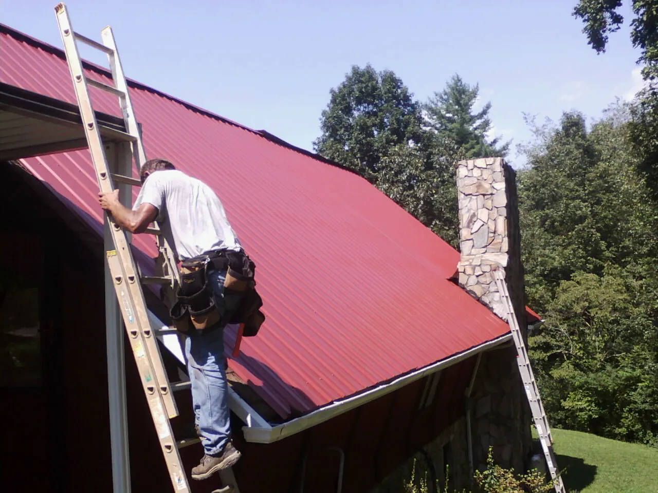 Roofer on a ladder beside a red metal roof and stone chimney — Soffit & Fascia Repair by Roofing Brothers in Luray